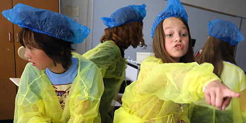 Students in yellow lab coats and blue caps pointing